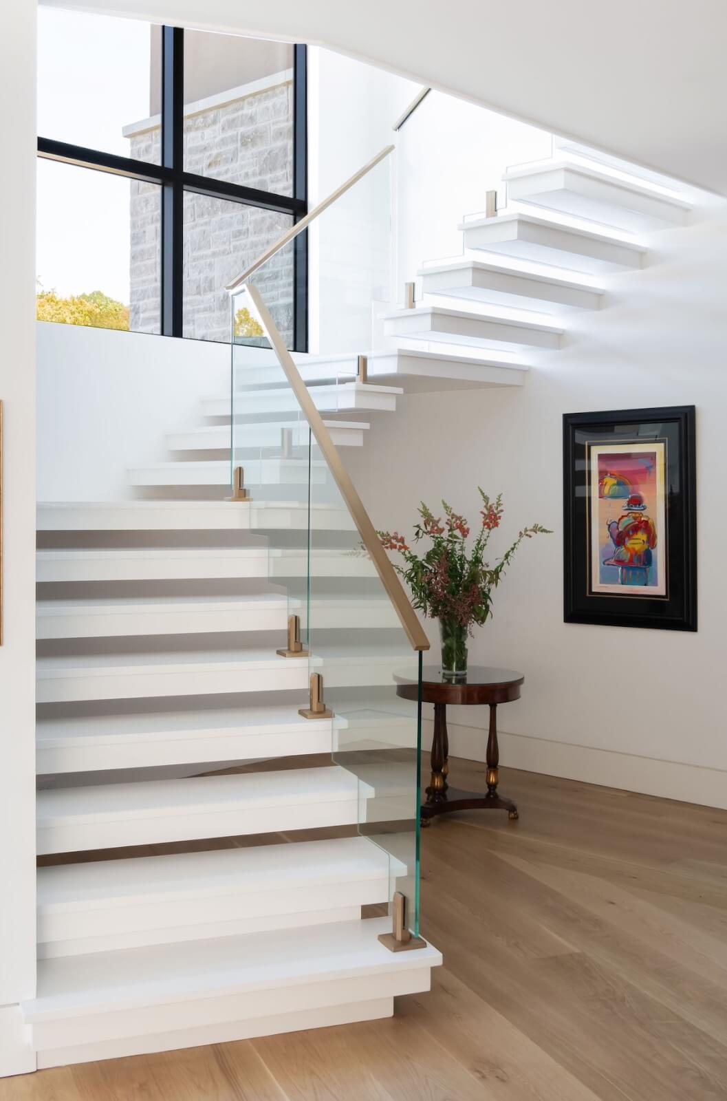 2 Homes by Beth Haley Design: A Study in Style & Contrast - 8 Modern interior by Beth Haley Design featuring a white floating staircase, glass railing, and wooden accents. Large windows and colorful framed artwork adorn the walls in this stunning home. A small table with flowers sits nearby, enhancing the elegant ambiance.