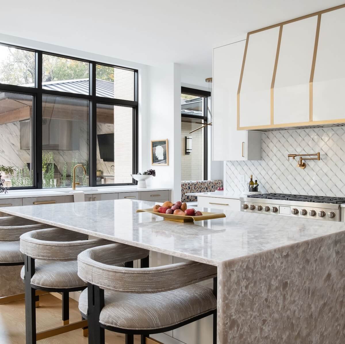 2 Homes by Beth Haley Design: A Study in Style & Contrast - 2 This modern kitchen by Beth Haley Design includes a large marble island, cushioned stools, a fruit tray, gas stove, brass faucet, and large windows. The white walls and herringbone backsplash enhance the space's stunning charm.