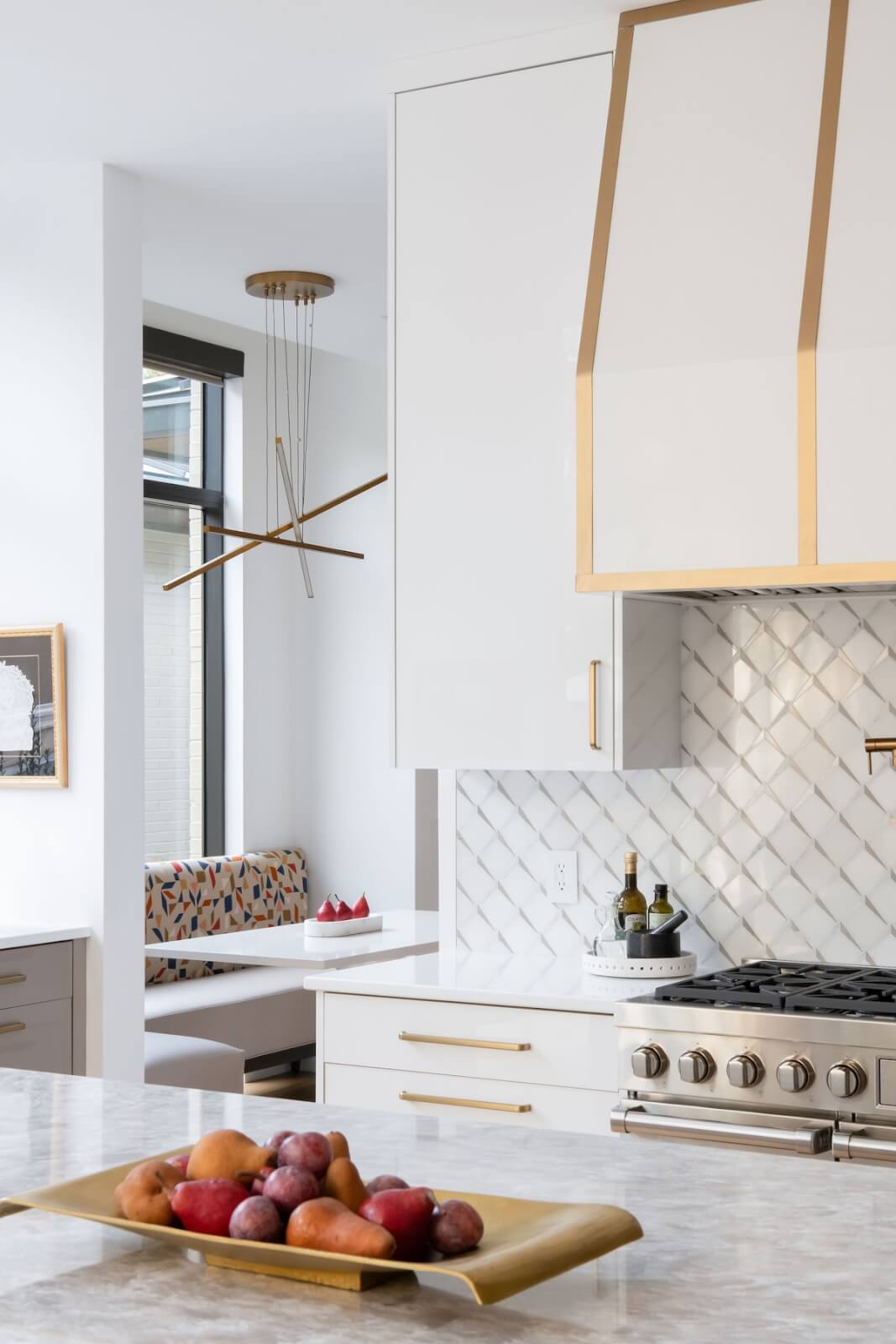 2 Homes by Beth Haley Design: A Study in Style & Contrast - 9 A modern kitchen with white cabinets and gold accents by Beth Haley Design. A gas stove is under a herringbone-patterned backsplash. A fruit tray on a marble island adds charm, while a dining nook and geometric light fixture reflect a blend of old and new elegance.