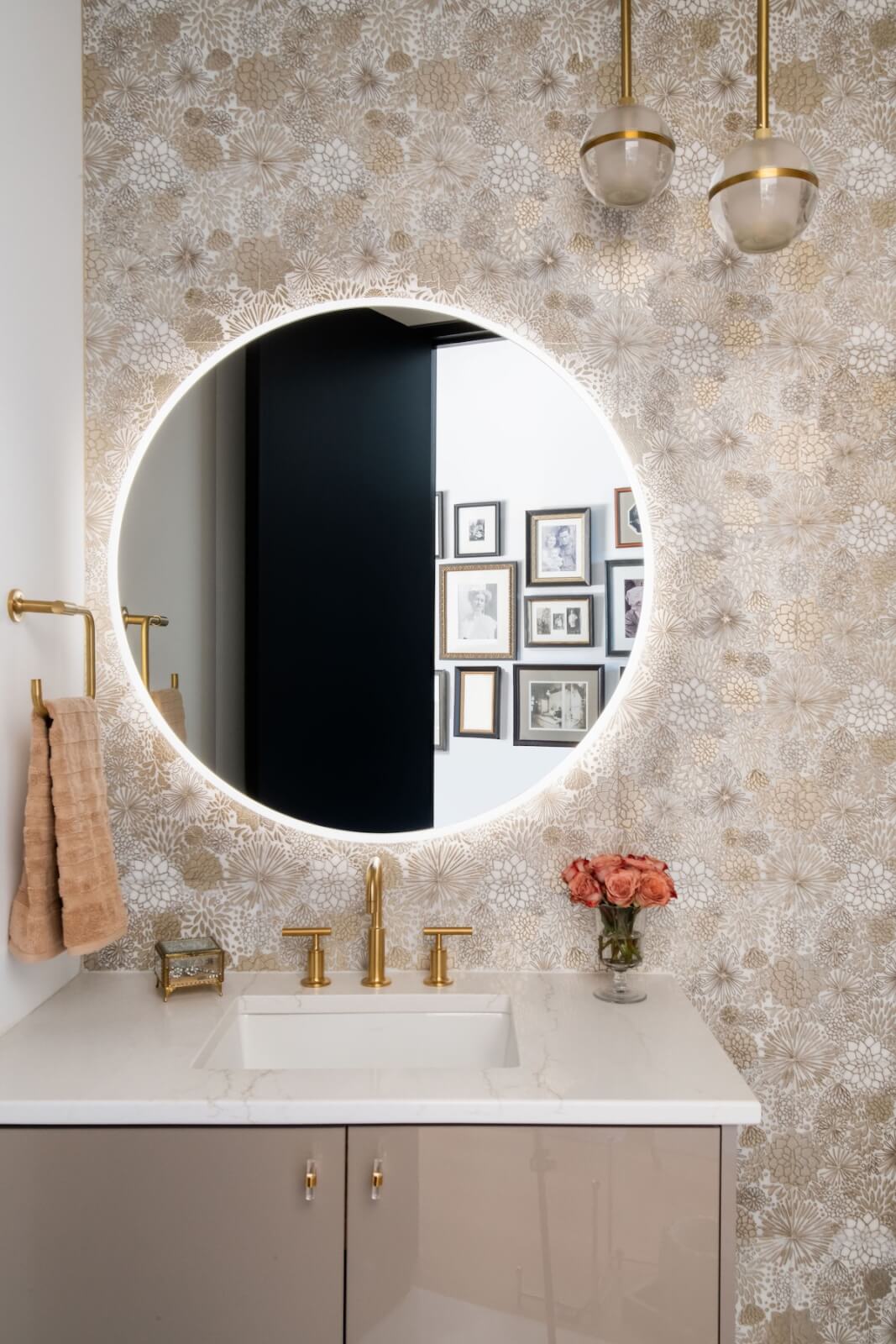 2 Homes by Beth Haley Design: A Study in Style & Contrast - 5 A bathroom blends old and new with a round, backlit mirror above the sink. Gold fixtures complement a beige towel and flowers on the countertop. The wall's floral pattern adds charm, while framed pictures in the background complete this creation by Beth Haley Design.