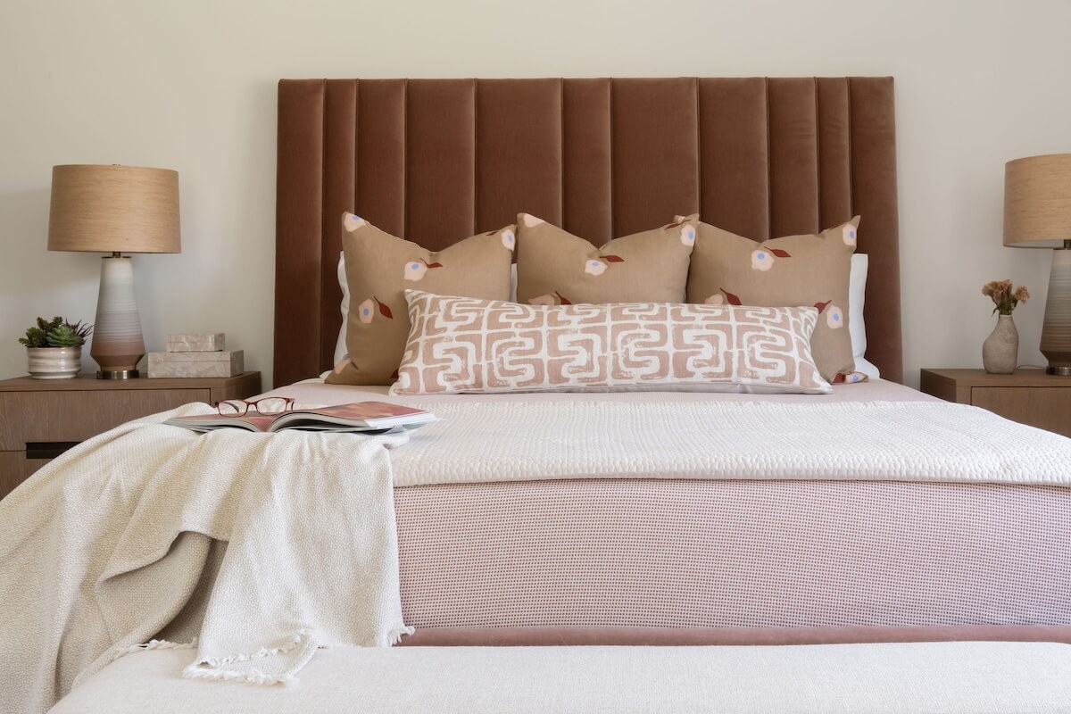 2 Homes by Beth Haley Design: A Study in Style & Contrast - 4 A neatly made bed with multiple cushions is flanked by two identical wooden nightstands, each with a lamp. An open book and throw blanket rest on the foot of this space.