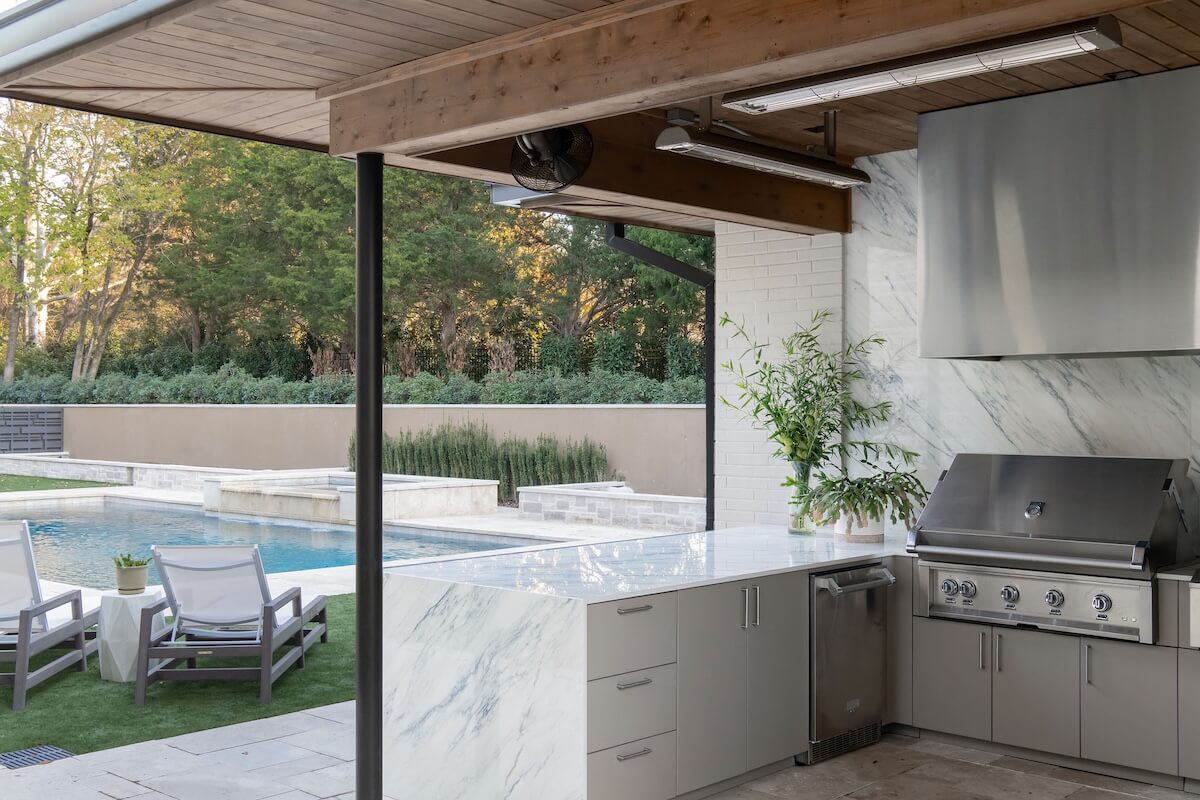 2 Homes by Beth Haley Design: A Study in Style & Contrast - 3 This stunning outdoor kitchen by Beth Haley Design features a stainless steel grill, marble countertops, and a potted plant. It elegantly overlooks a pool area with patio chairs and lush greenery.
