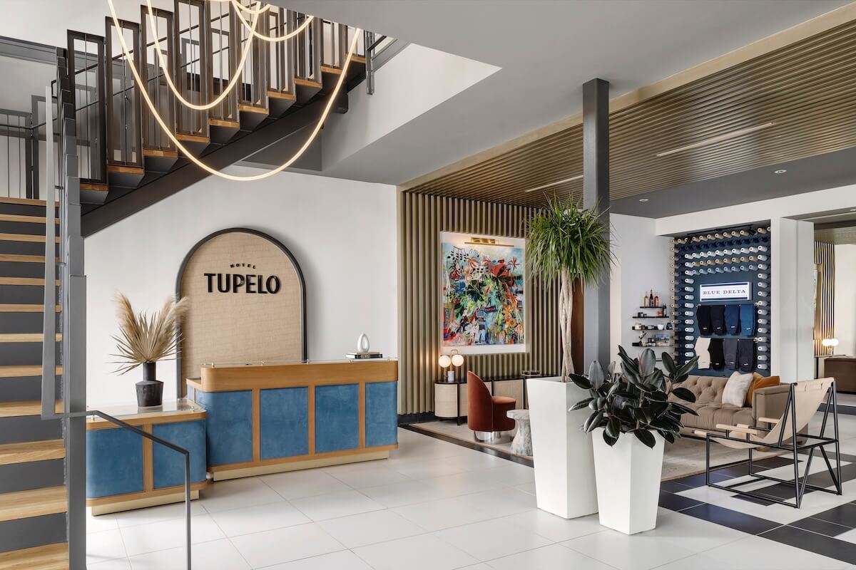Modern hotel lobby in Tupelo MS with a blue and wood reception desk labeled "Tupelo," contemporary furniture, large plants, and decorative art—perfect for trip planning or starting a memorable girls' trip.