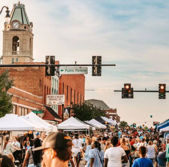 The Best Festivals in the South - 11 A large crowd attends one of the best festivals in the South, enjoying an outdoor street festival with tents and vendor booths, under traffic lights and a clock tower in the background on a sunny day.