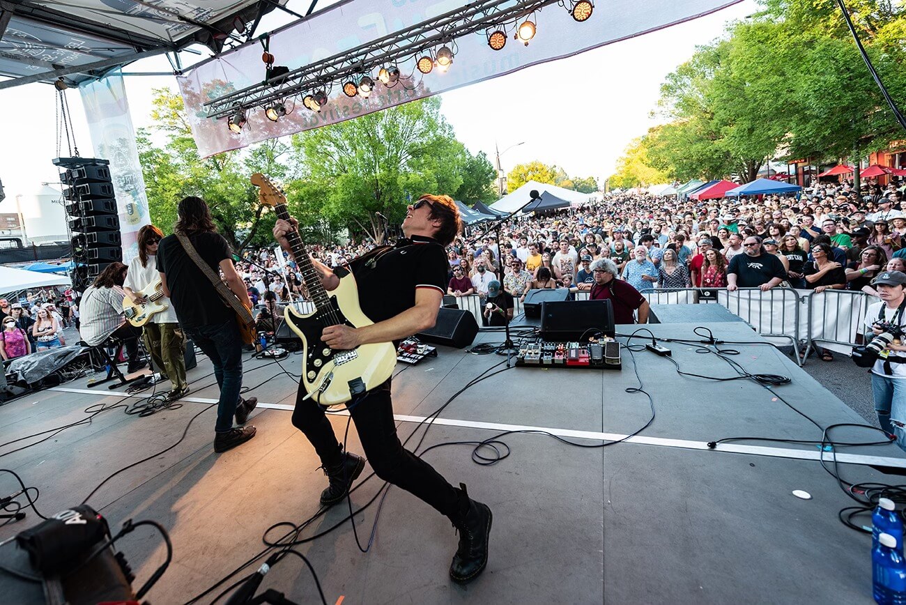 The Best Festivals in the South - 13 The band played energetically on stage at one of the South's best festivals, captivating a large crowd in broad daylight. Trees and tents formed a vibrant backdrop as festival-goers soaked in the lively atmosphere.