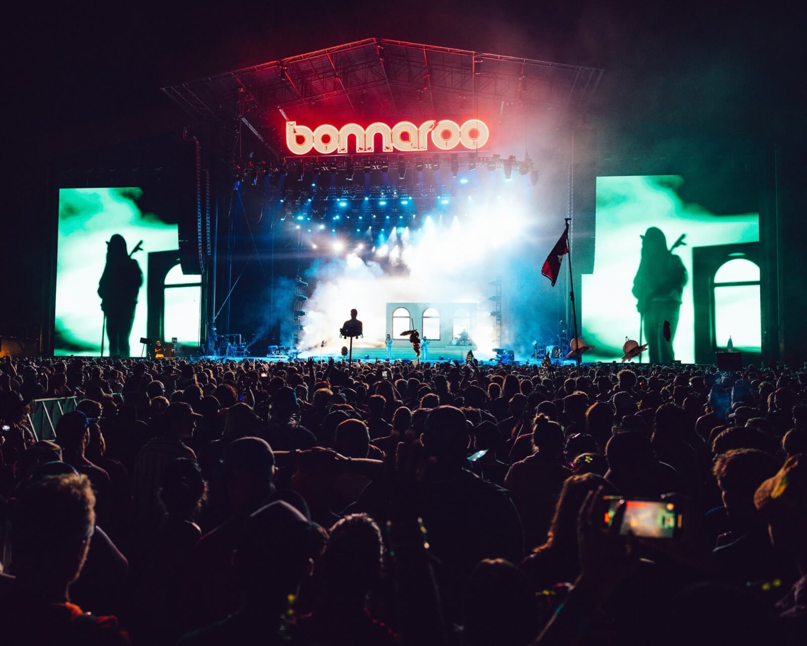 The Best Festivals in the South - 12 A lively crowd gathers beneath the glowing "Bonnaroo" sign at a nighttime outdoor concert in the South. The festival's vibrant energy is captured on large screens, where silhouetted figures dance against a colorful backdrop.