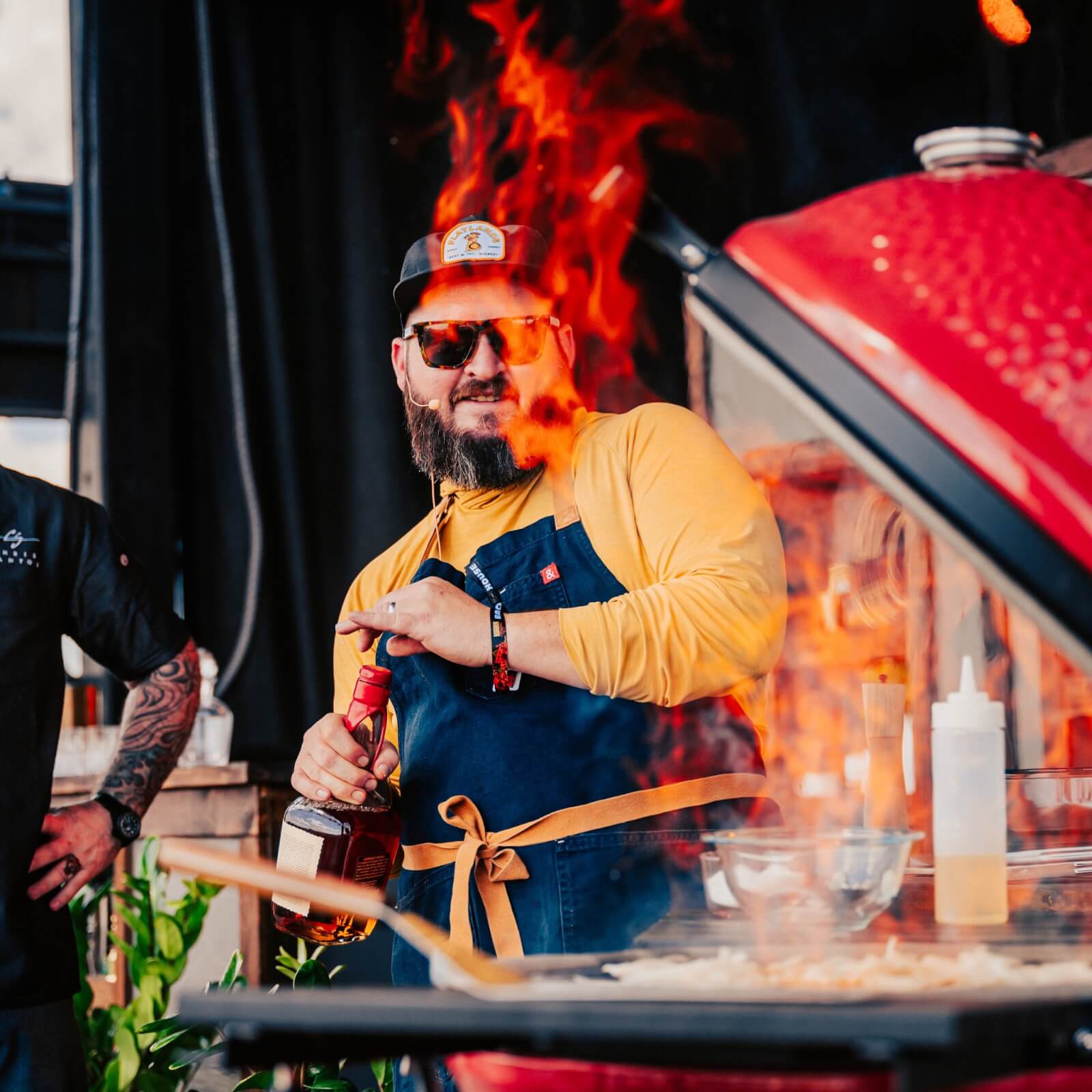 The Best Festivals in the South - 17 At a vibrant Southern festival, a bearded chef wearing sunglasses and a cap showcases his grilling skills. Flames leap from the grill as he expertly wields a bottle, captivating onlookers with his fiery culinary prowess.