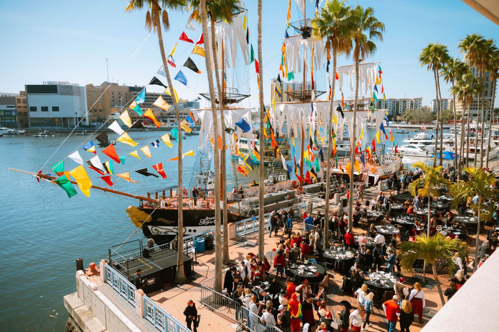 The Best Festivals in the South - 2 Colorful flags flutter on tall ships at a lively waterfront festival in the South, as a crowd mingles under the shade of swaying palm trees.