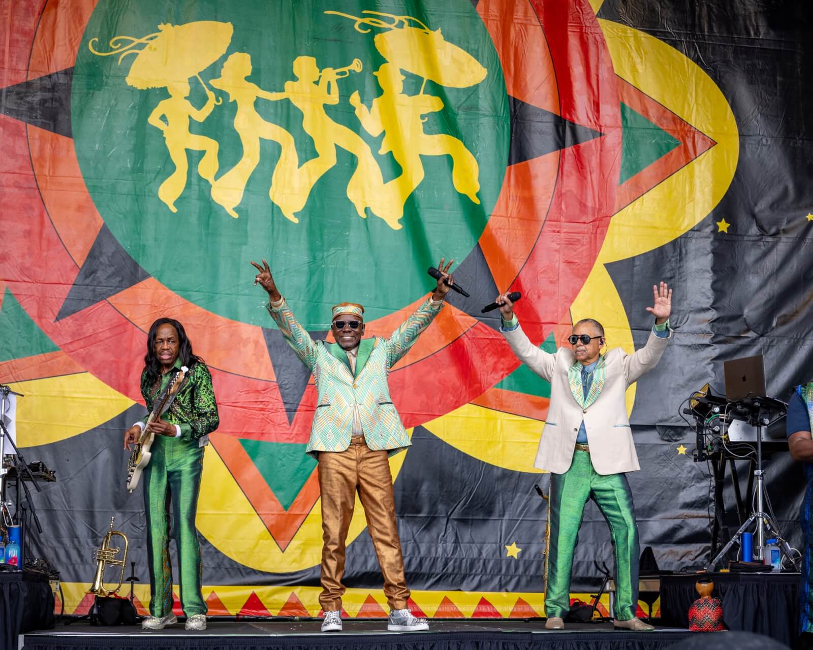 The Best Festivals in the South - 5 Three performers on stage in colorful suits, with a vibrant backdrop featuring a star and trumpet players, capture the lively essence of Southern festivals. Two hold microphones while the third strums a guitar, enchanting the audience with their melodies.