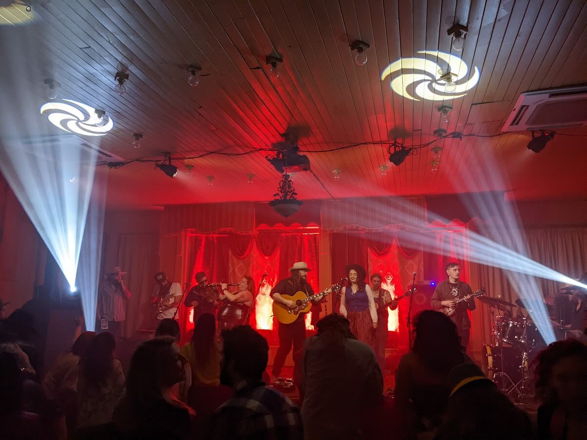 The Best Festivals in the South - 1 A band performs on stage with vibrant lighting. The red backdrop highlights musicians playing guitars and violins, while the audience stands in silhouette, captivated by the performance.