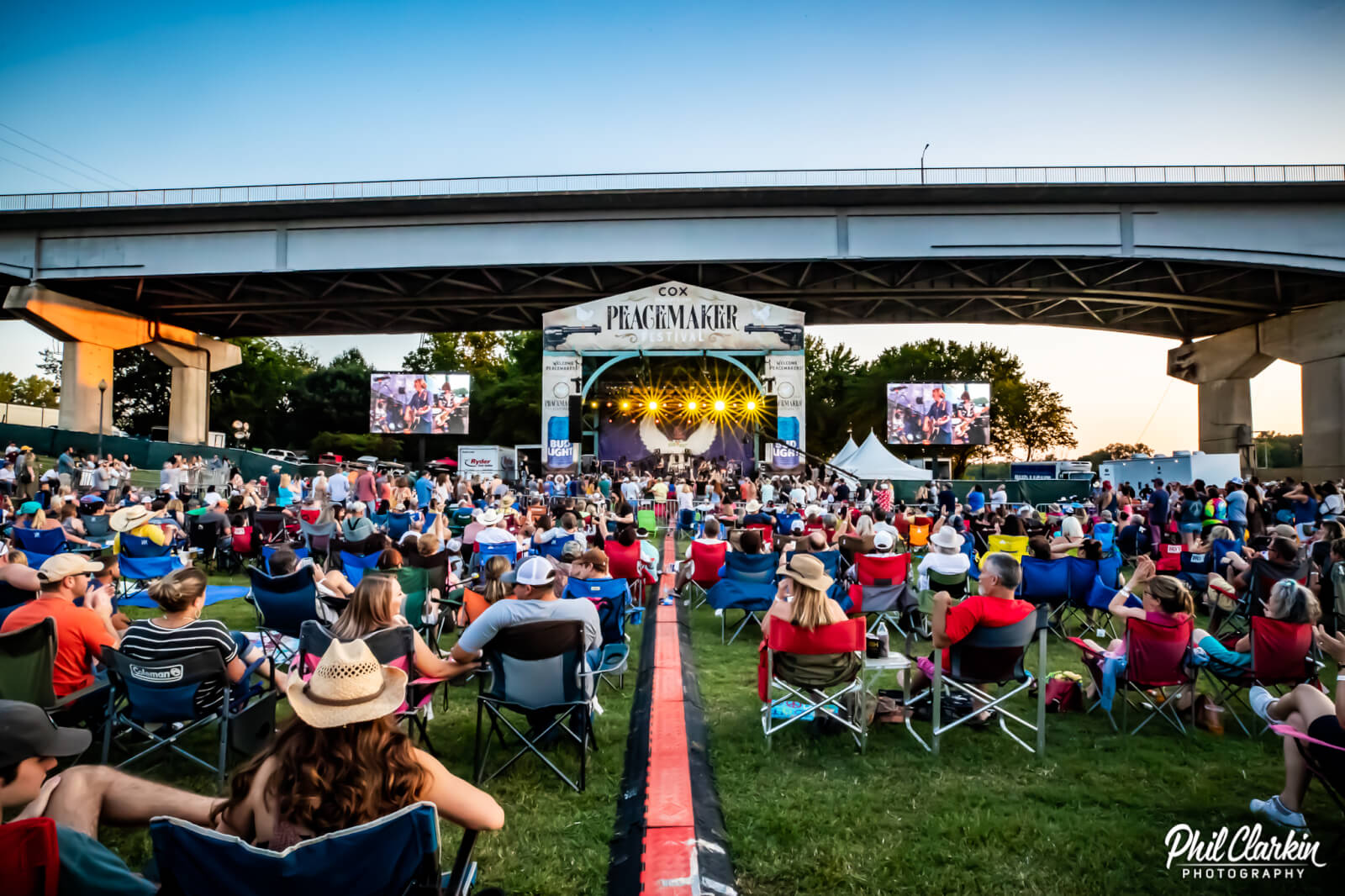The Best Festivals in the South - 18 A large crowd sits on lawn chairs facing an outdoor concert stage under a highway overpass at sunset during the Peacemaker Festival, one of the best festivals in the South.