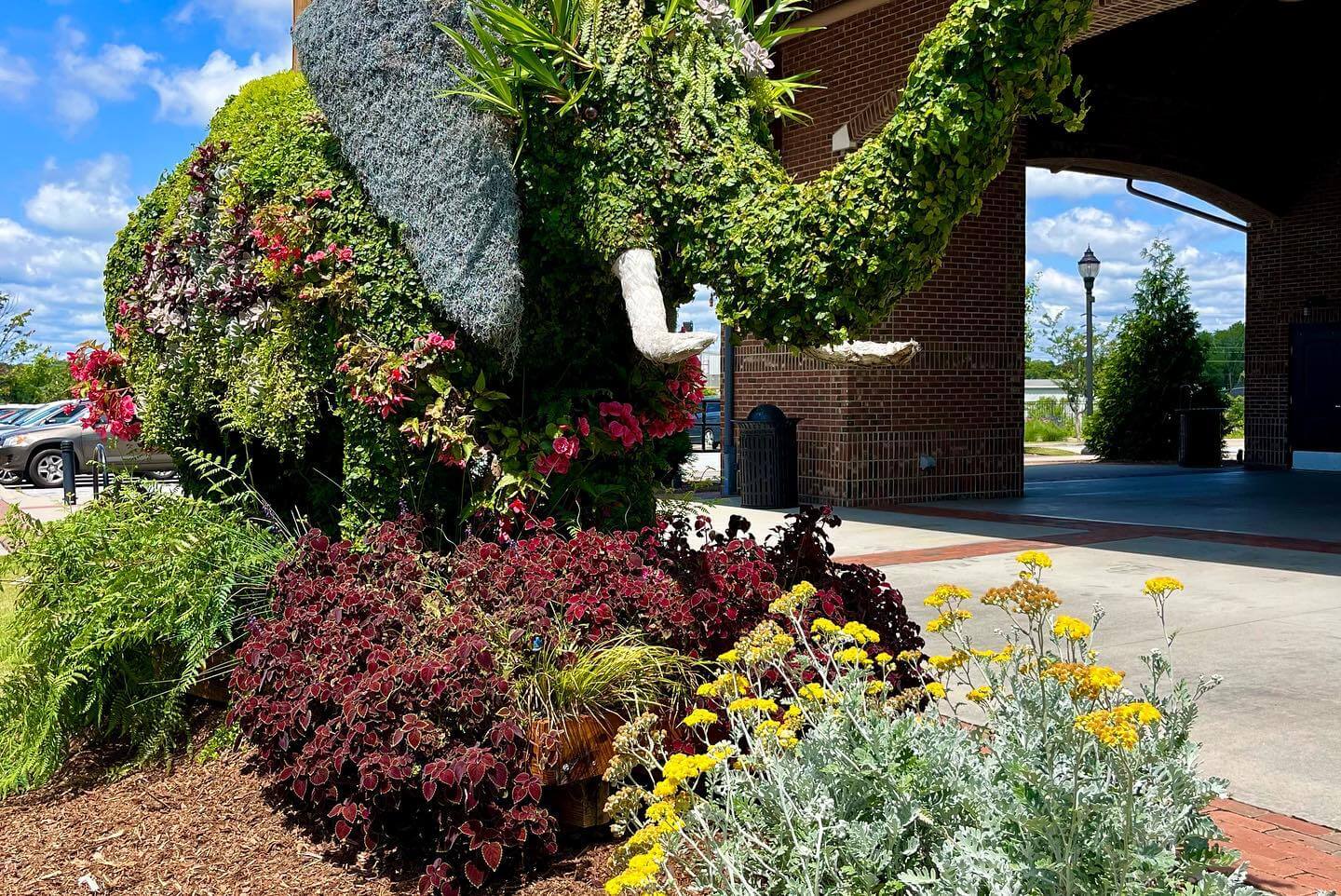 The Best Festivals in the South - 9 A topiary elephant covered in greenery and flowers stands proudly next to a brick building, embracing the festive spirit of the South. Surrounded by landscaped plants, including vibrant yellow and red flowers, it basks under a partly cloudy sky.