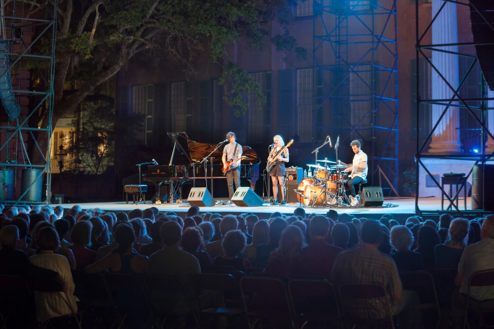 The Best Festivals in the South - 7 A quartet performs on an outdoor stage with guitar, piano, saxophone, and drums during a lively festival in the South. The audience watches under dim lighting with scaffolding and trees in the background, soaking in the ambiance of this spirited event.