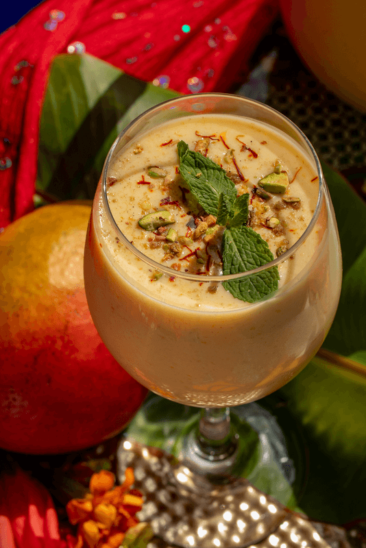 A glass of mango lassi garnished with chopped pistachios, saffron strands, and mint leaves sits on a table—an ideal refreshment after exploring the Best Trails or hiking South, with a mango and decorative fabric in the background.