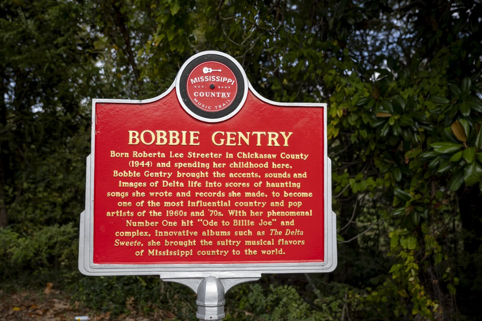 The red historical marker in Mississippi celebrates Bobbie Gentry's Southern roots and influence as a country music legend, highlighting her iconic song "Ode to Billie Joe." 