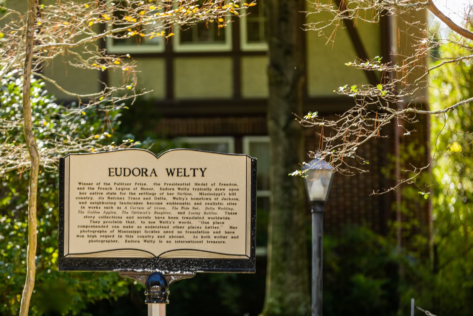 A historical marker for Eudora Welty stands in front of a charming house, nestled among trees and a lamp post, detailing her literary achievements and awards. This spot is a highlight on Southern literary trails, celebrating the best of regional storytelling.