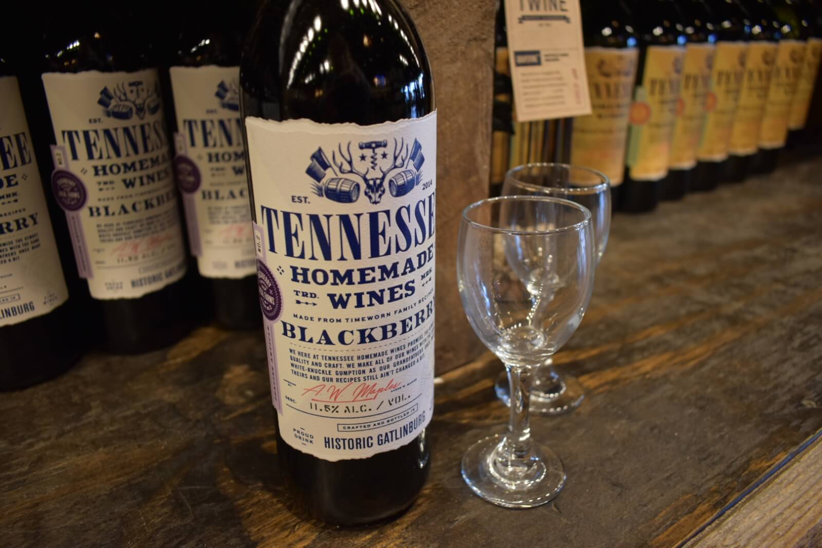 A bottle of Tennessee Homemade Wines Blackberry wine is displayed on a wooden surface next to two empty wine glasses, with other wine bottles in the background.