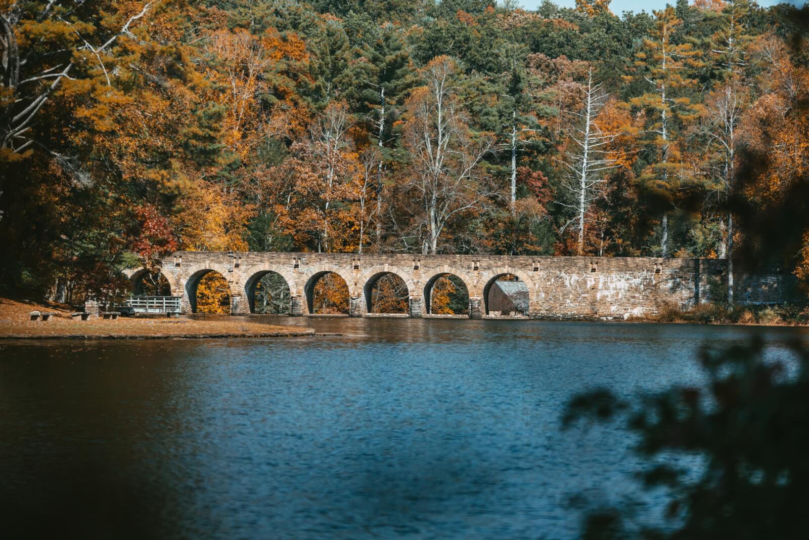 Stone bridge spanning across lake, with fall foliage in the background.