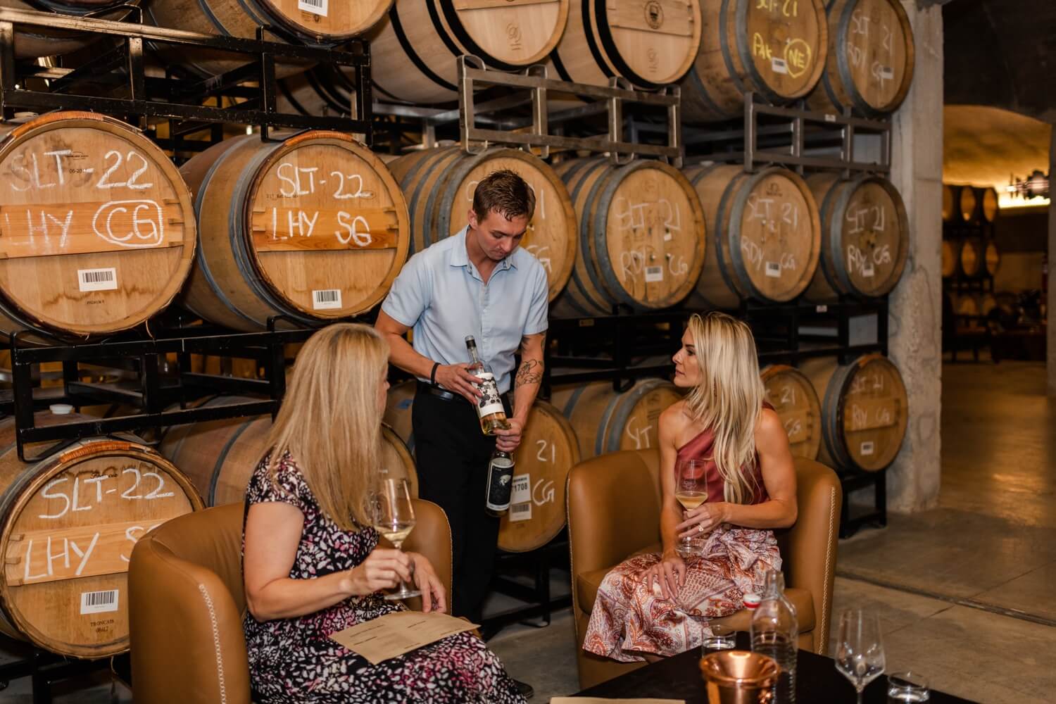 A man pours wine for two women seated in a cellar with stacked barrels in the background, celebrating a well-earned rest after exploring top trails and scenic hikes in the South.