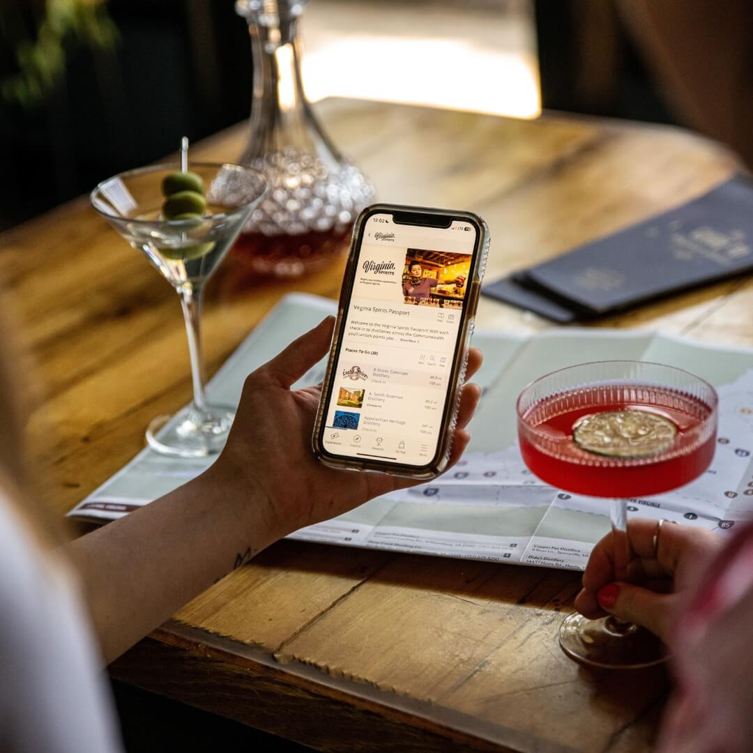 A person holding a smartphone displaying a wine app, seated at a wooden table with cocktails, a wine decanter, and a folded map of top trails and hikes in the South.