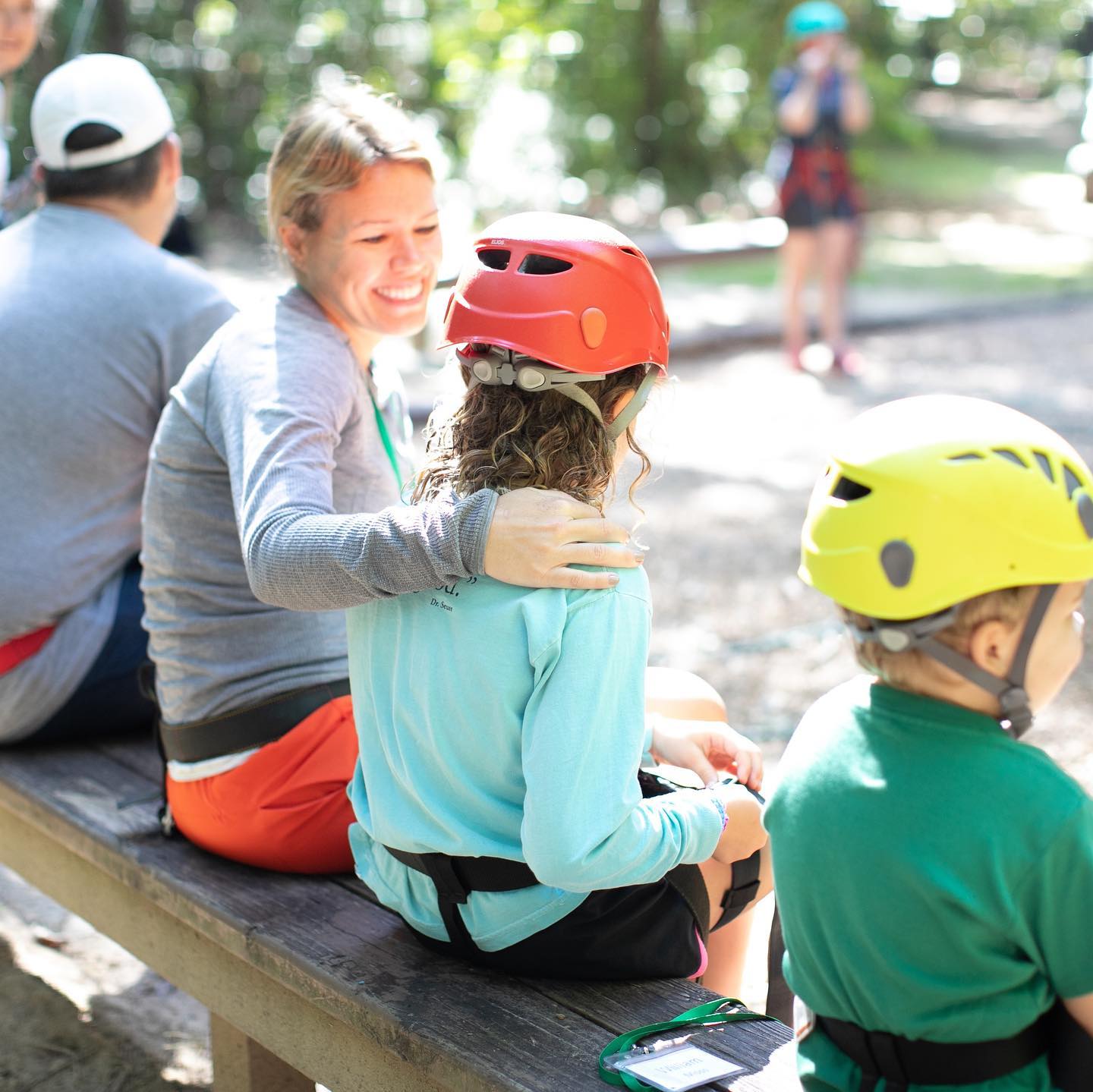 A woman sits on a bench during summer camp, smiling as she places her hand on a child's back. The child, wearing a red helmet, stands next to another in a yellow helmet, all enjoying summer activities with the group outdoors.