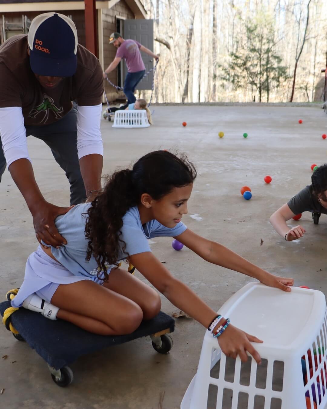 A child on a wheeled platform reaches for a laundry basket, assisted by an adult during Family Camp. Other children and colorful balls, reminiscent of a vibrant summer day, are visible in the background.