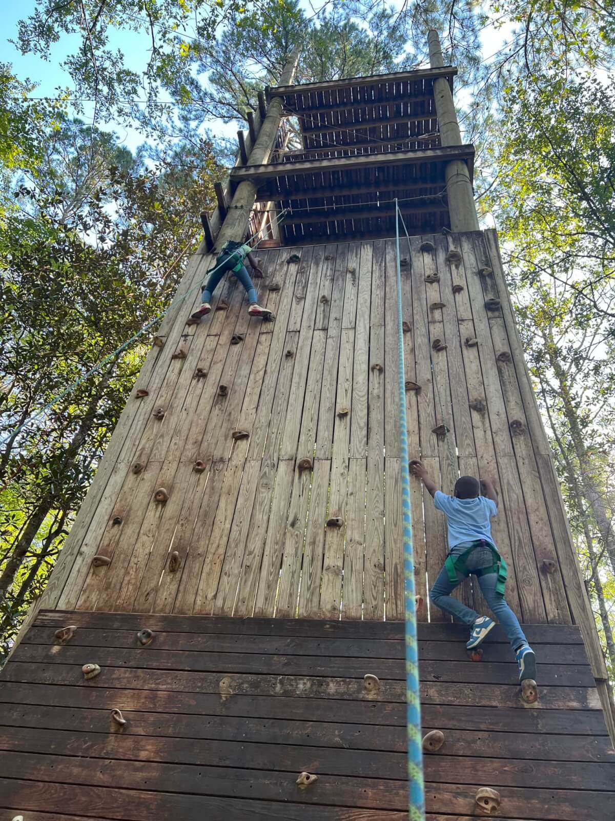 Two children enjoy a Family Camp adventure, climbing a wooden rock wall outdoors amidst towering trees, secured with a safety rope.
