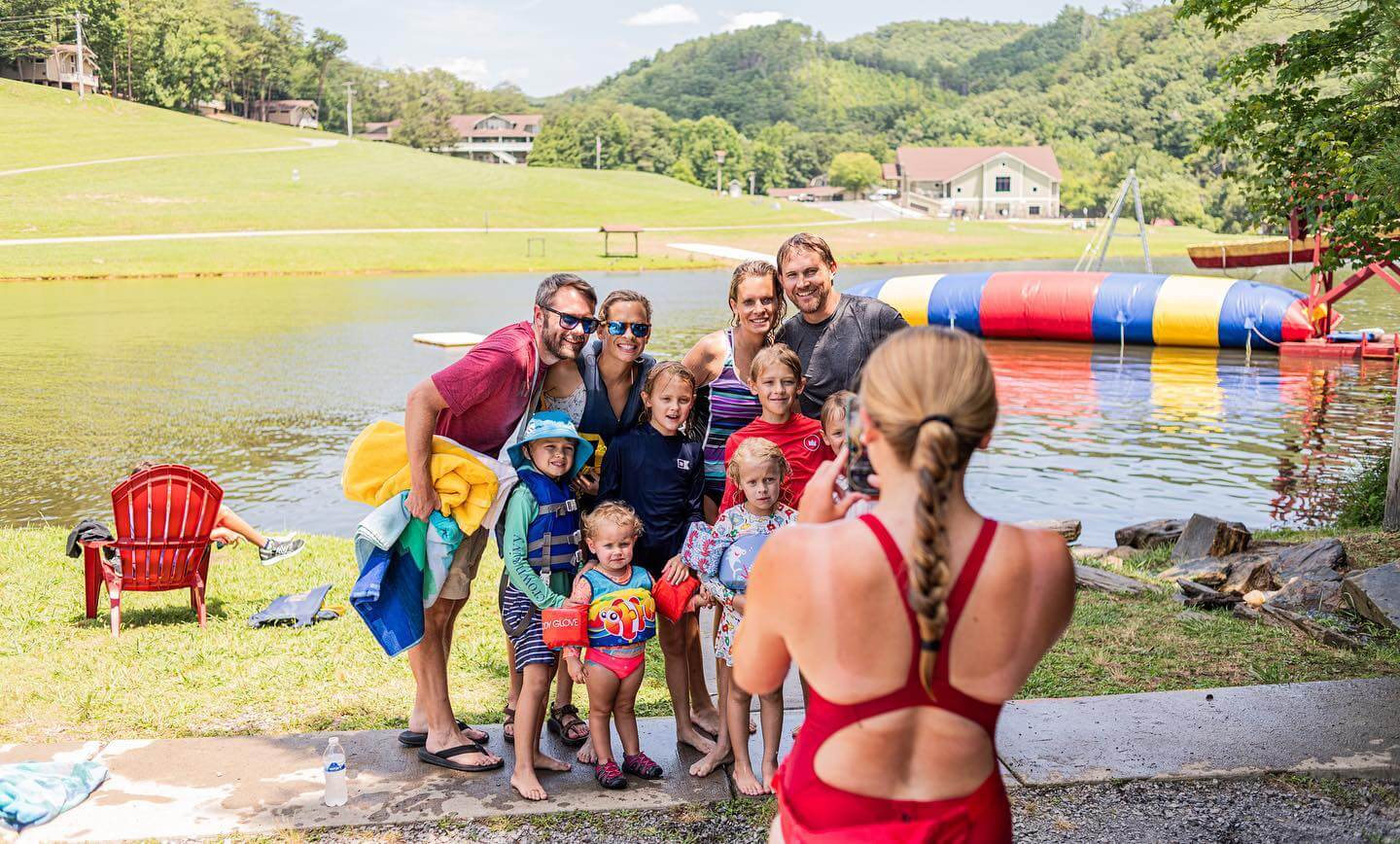A group of people, including children in swimwear, pose for a photo near a lake during their Family Camp. A colorful inflatable adds to the summer fun in the background as a person in a red swimsuit takes the picture.