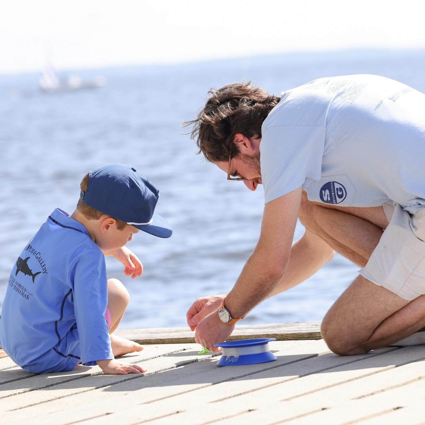 An adult and child enjoy a serene moment at family camp, sitting casually on the dock by the water. Together, they examine a small object, with the child sporting a blue cap and shirt.