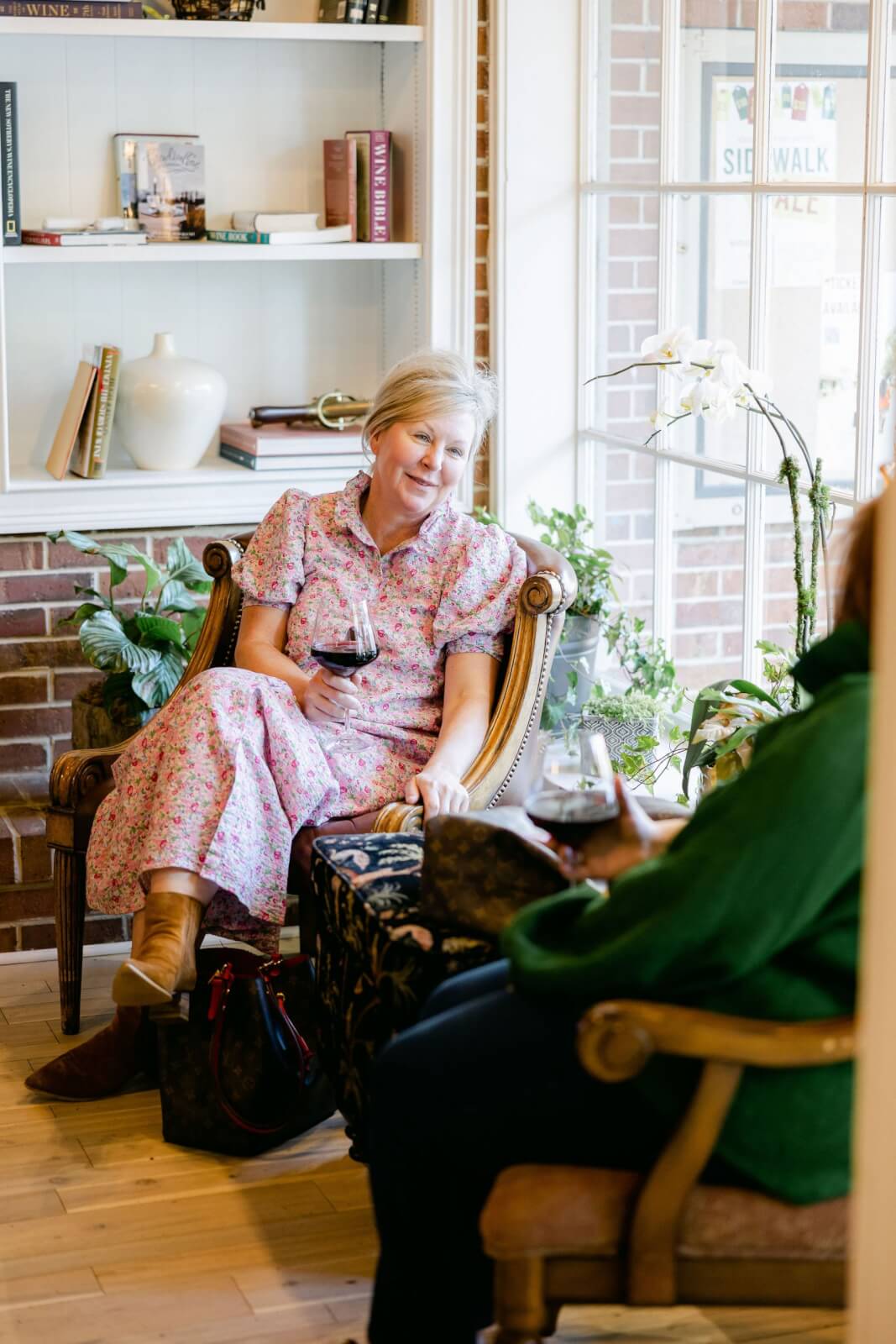 Georgia's 6 Best Girlfriends' Weekend Getaways - 17 Two people lounging in a cozy Georgia retreat, enjoying glasses of wine amidst lush plants and shelves filled with books. One wears a floral dress, the other a green top.