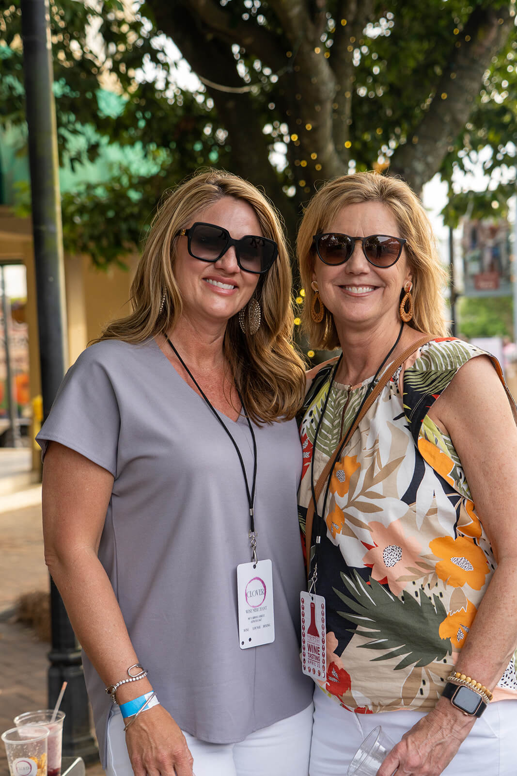 Georgia's 6 Best Girlfriends' Weekend Getaways - 18 Two women wearing sunglasses and name tags are smiling in front of a tree adorned with string lights, embodying the perfect vibe for girlfriend getaways. Casually dressed, one sports a floral top while the other opts for a gray shirt.