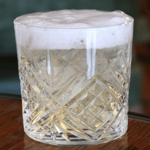 A clear, patterned glass filled with a fizzy, pale beverage topped with foam, placed on a wooden surface.