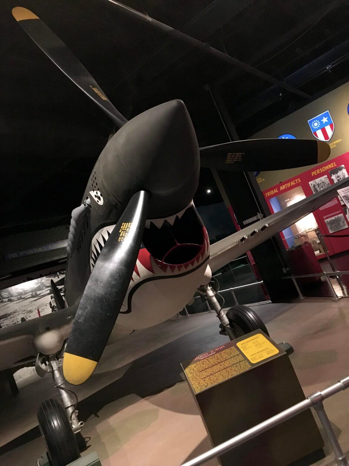 15+ Top Trails in the South for History, Hikes & Hometown Flavor - 1 World War II fighter plane with shark mouth nose art displayed in a museum exhibit in the South, with informational signs and photos in the background.