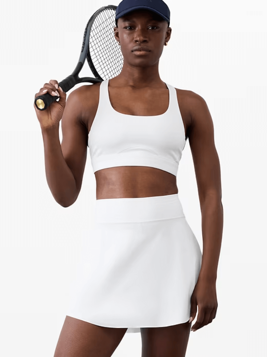 A person in golf attire—a white sports bra and skirt—holds a tennis racket over their shoulder, wearing a navy cap, standing against a plain background, offering inspiration on what to wear golfing with sporty flair.