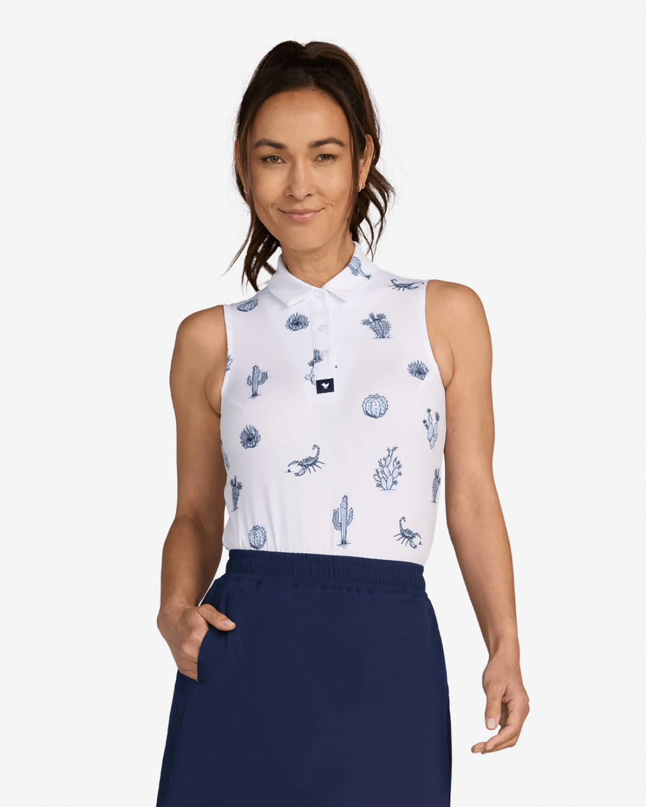 A woman wearing a sleeveless white collared shirt with blue desert-themed prints and a navy skirt, standing with one hand in her pocket, facing forward—showcasing stylish golf attire perfect for those wondering what to wear golfing.
