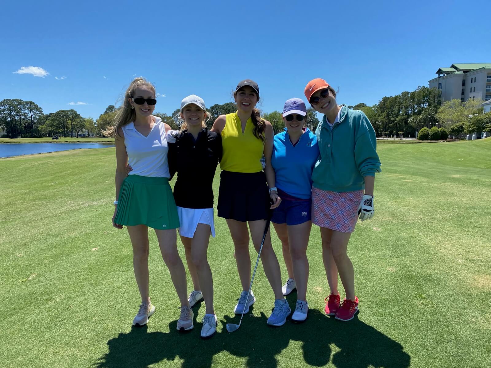 Five women stand together on a golf course in sunny weather, dressed in stylish golfing attire, smiling at the camera with a building and trees in the background.