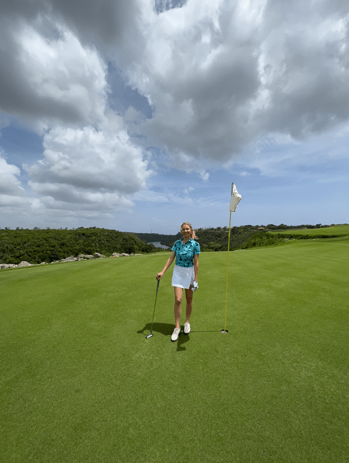 A woman stands on a golf course next to a flagstick, dressed in stylish golf attire, holding a golf club with green grass and a cloudy sky in the background.