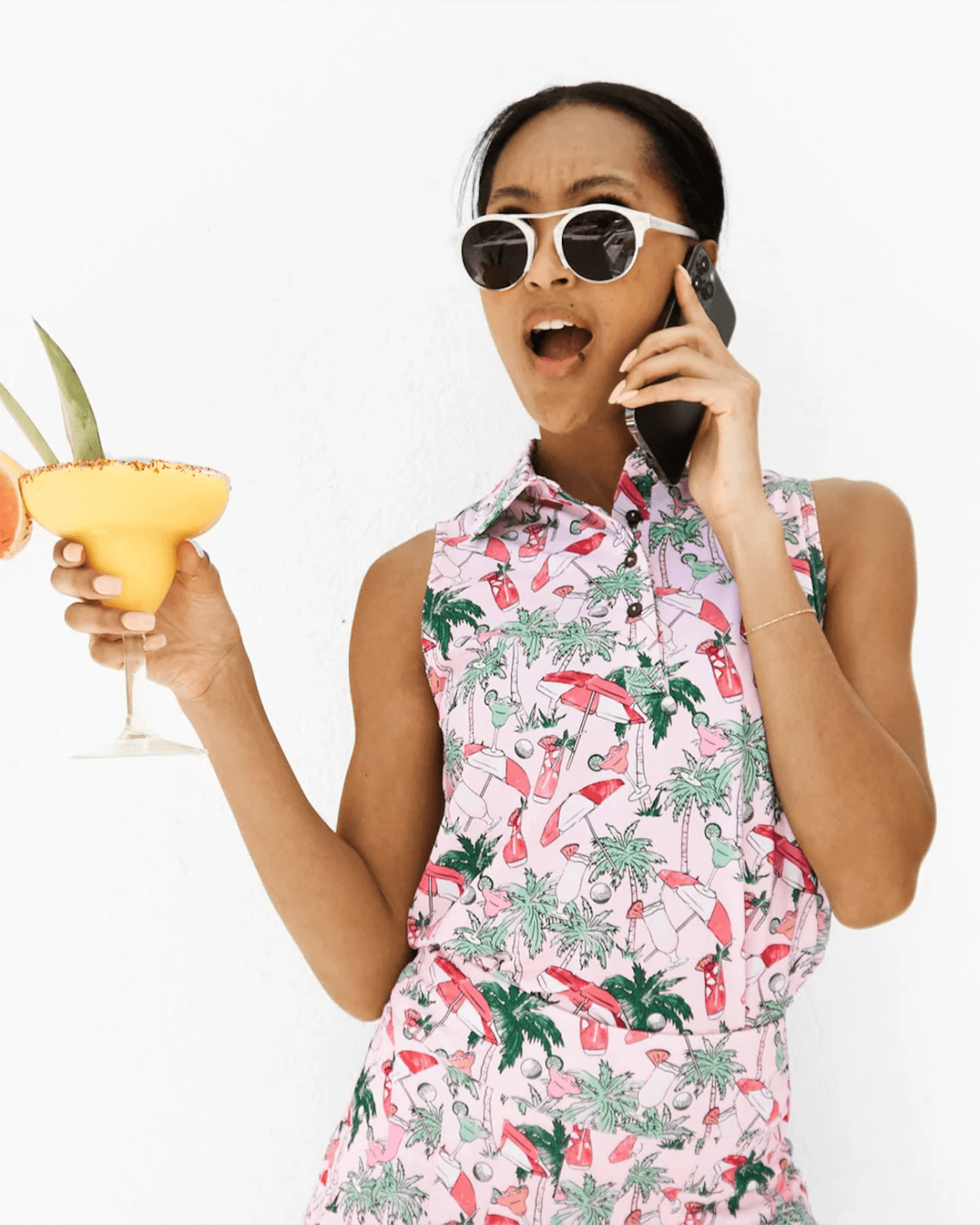 Woman wearing sunglasses and a colorful, golf-inspired sleeveless outfit holds a yellow cocktail and talks on the phone against a plain white background—a chic spin on golf and court outfits.