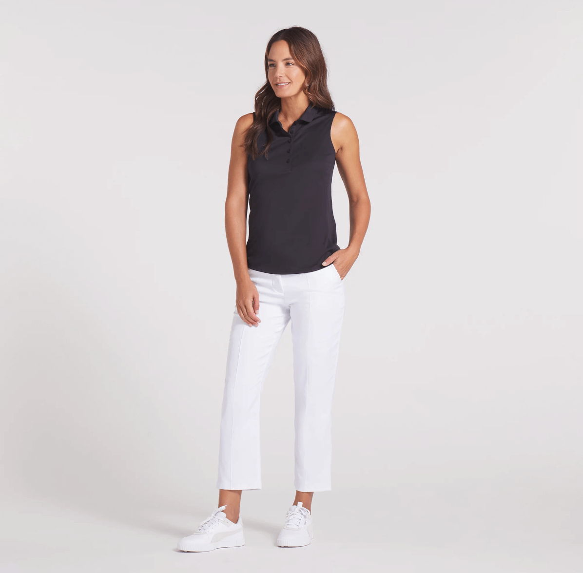 A woman stands against a plain background wearing a black sleeveless top, white cropped pants, and white sneakers—an effortlessly chic choice for golf attire or a stylish court outfit, with one hand casually placed in her pocket.