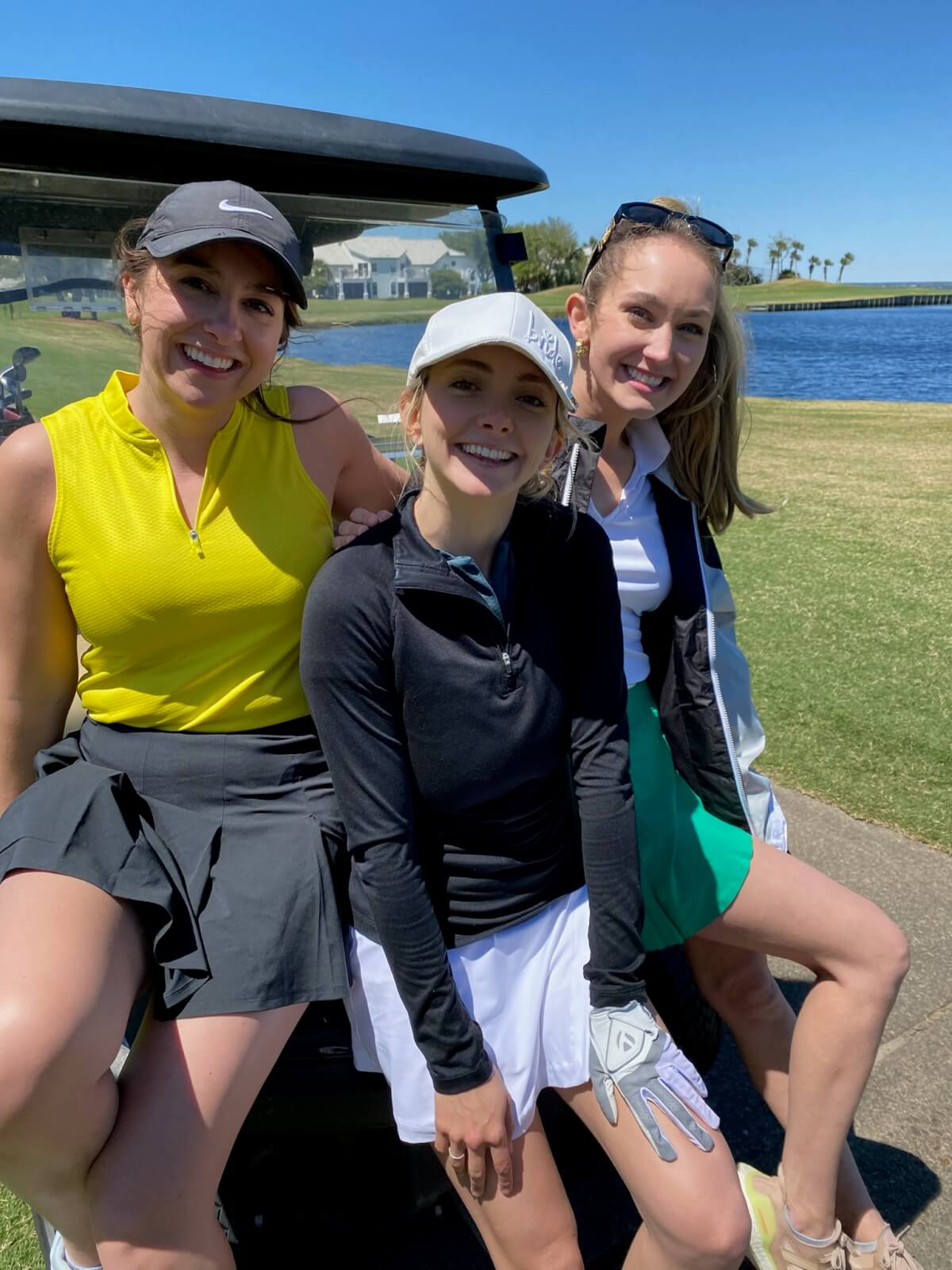 Three women showcase stylish golf attire as they sit and stand by a golf cart on a sunny day, with a scenic golf course, water, and palm trees in the background—offering inspiration for what to wear golfing.