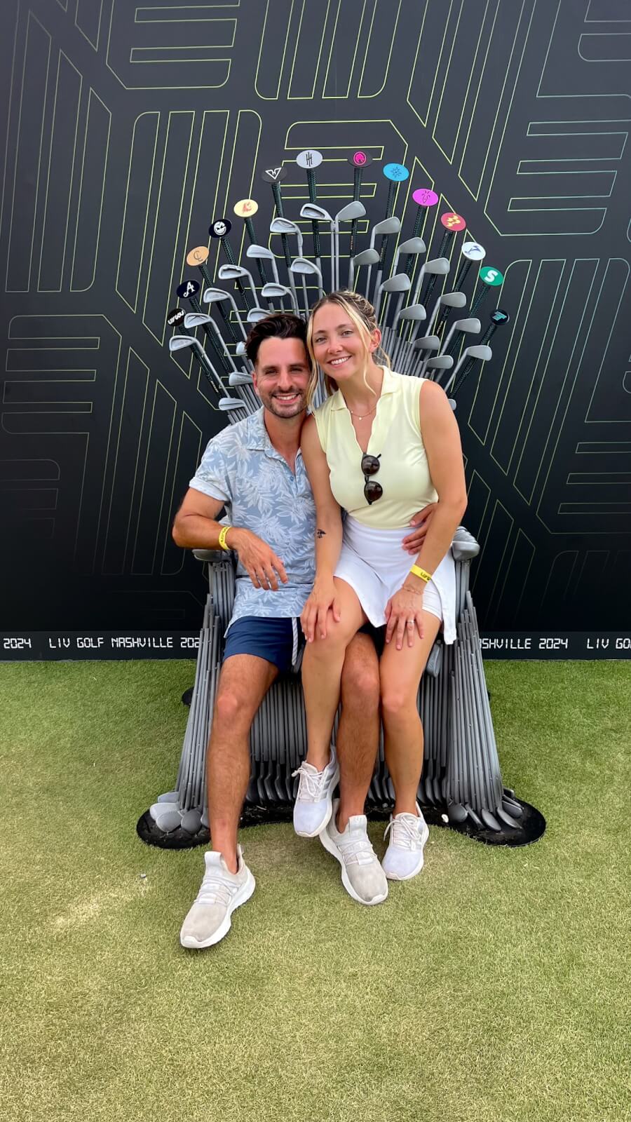 A man and woman in golf attire sit together on a chair made of golf clubs, smiling at the camera, with a geometric black backdrop behind them.