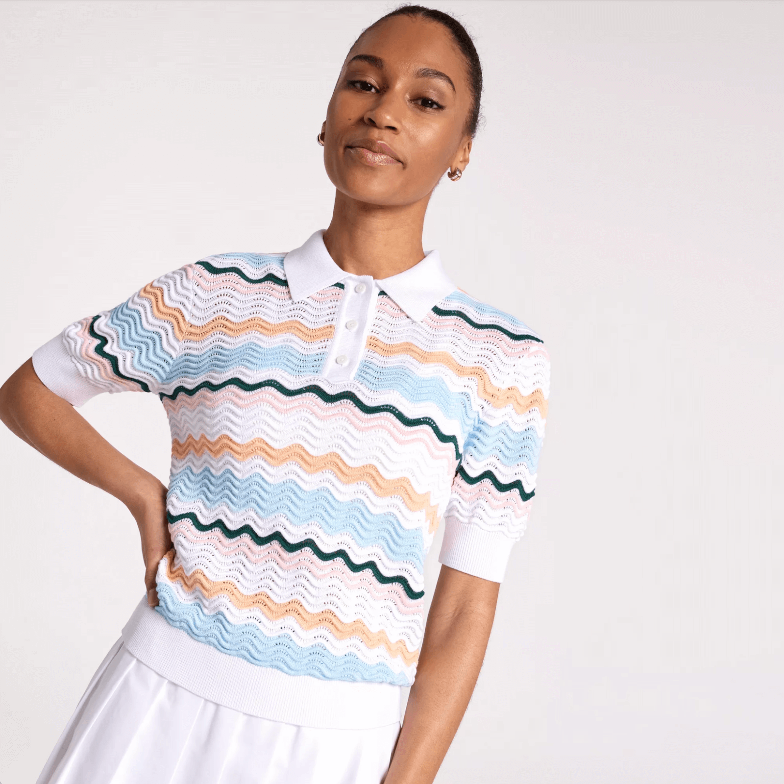 A woman wearing a white collared short-sleeve knit top with multicolored wavy stripes and a white skirt—perfect golf attire inspiration for those wondering what to wear golfing—stands against a plain light background.