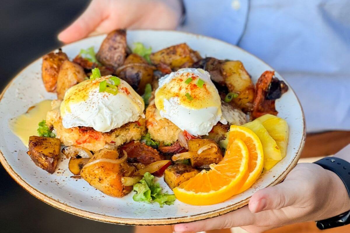A person holds a plate with two poached eggs on crab cakes, roasted potatoes, mixed vegetables, lettuce, and orange and pineapple slices—an ideal dish for Easter brunch in Birmingham.