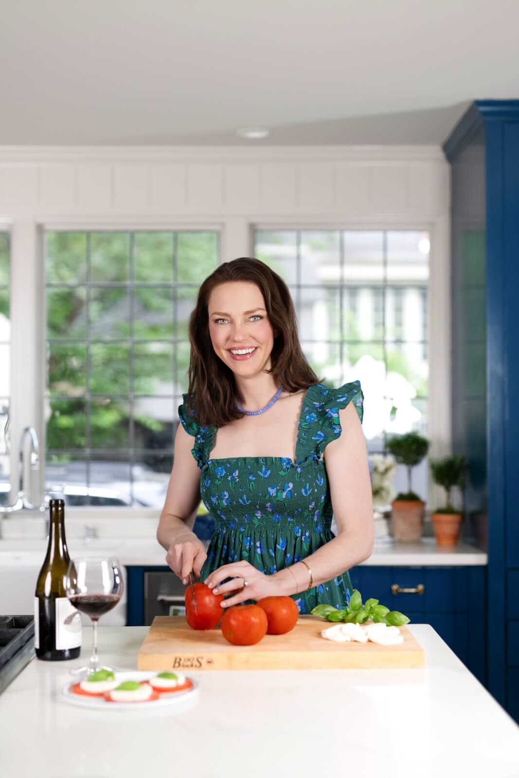 Dr. Julia Tauscher chops tomatoes on a wooden cutting board in a bright kitchen, enjoying her skincare routine. A glass of red wine, a wine bottle, and a plate of sliced vegetables are nearby.