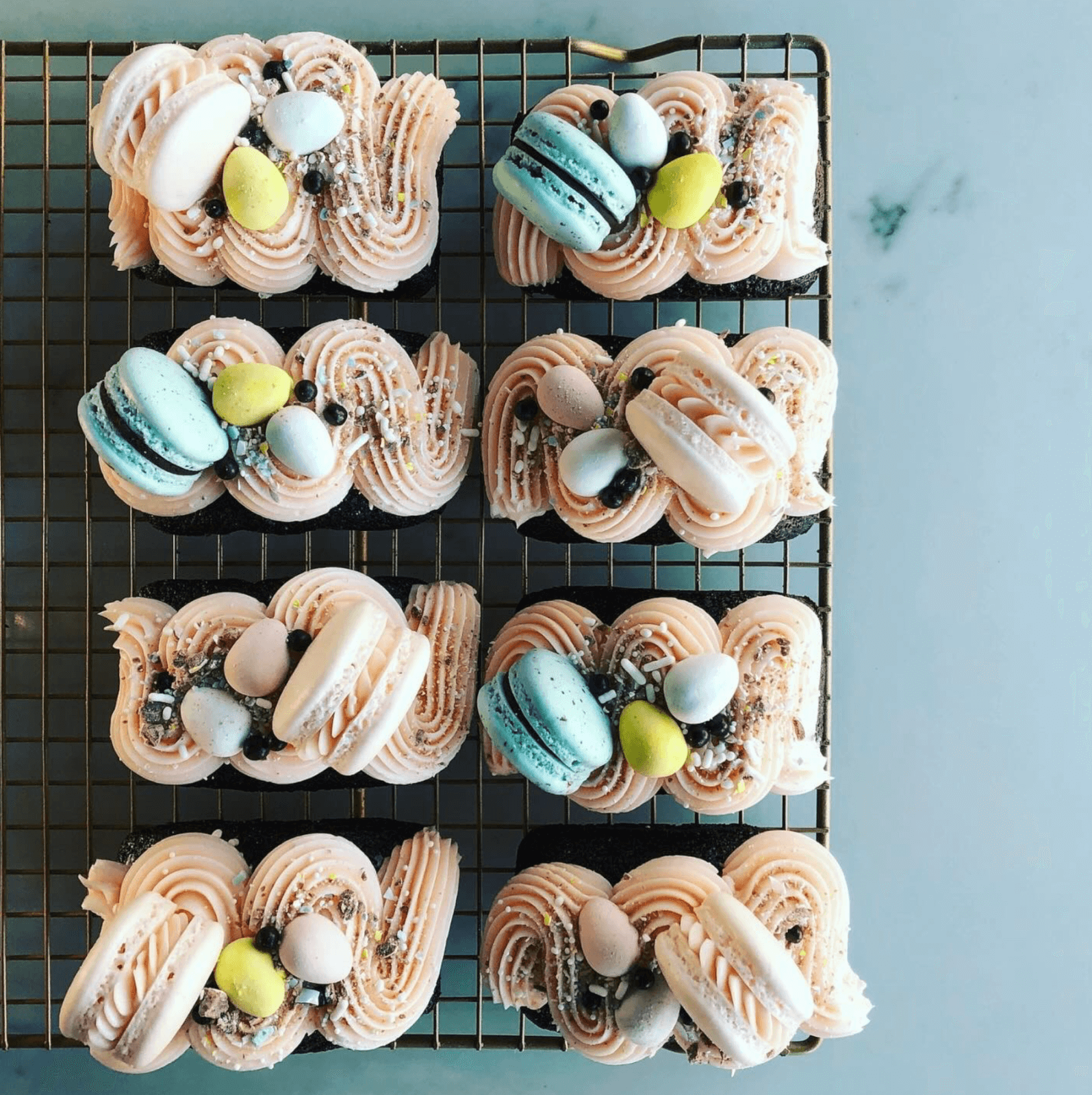 Nine rectangular chocolate cakes topped with pink swirled frosting, pastel macarons, and candy eggs are arranged on a wire cooling rack—perfect treats for celebrating new restaurants in Birmingham.