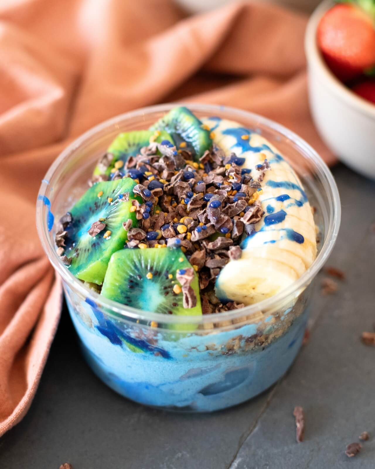 A clear bowl filled with blue smoothie, kiwi slices, banana slices, granola, cacao nibs, and blue drizzle sits on a gray surface with a peach napkin.