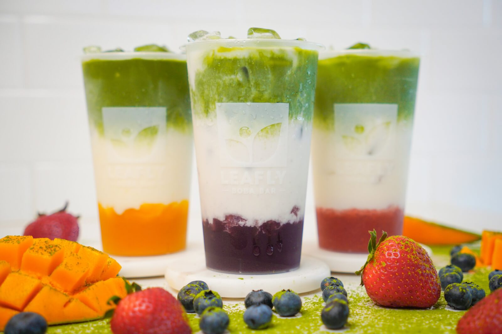 Three layered iced drinks in clear cups with fruit pieces and leaves, featuring vibrant gradients of green, white, and orange or purple—perfect refreshment found at trendy new restaurants in Birmingham, surrounded by fresh strawberries, mango, and blueberries.