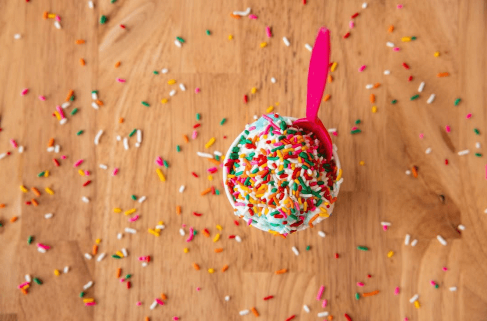 A cup of ice cream with rainbow sprinkles and a pink spoon sits on a wooden surface, showcasing the sweet side of Birmingham Dining, with sprinkles scattered around for an extra touch of delight.