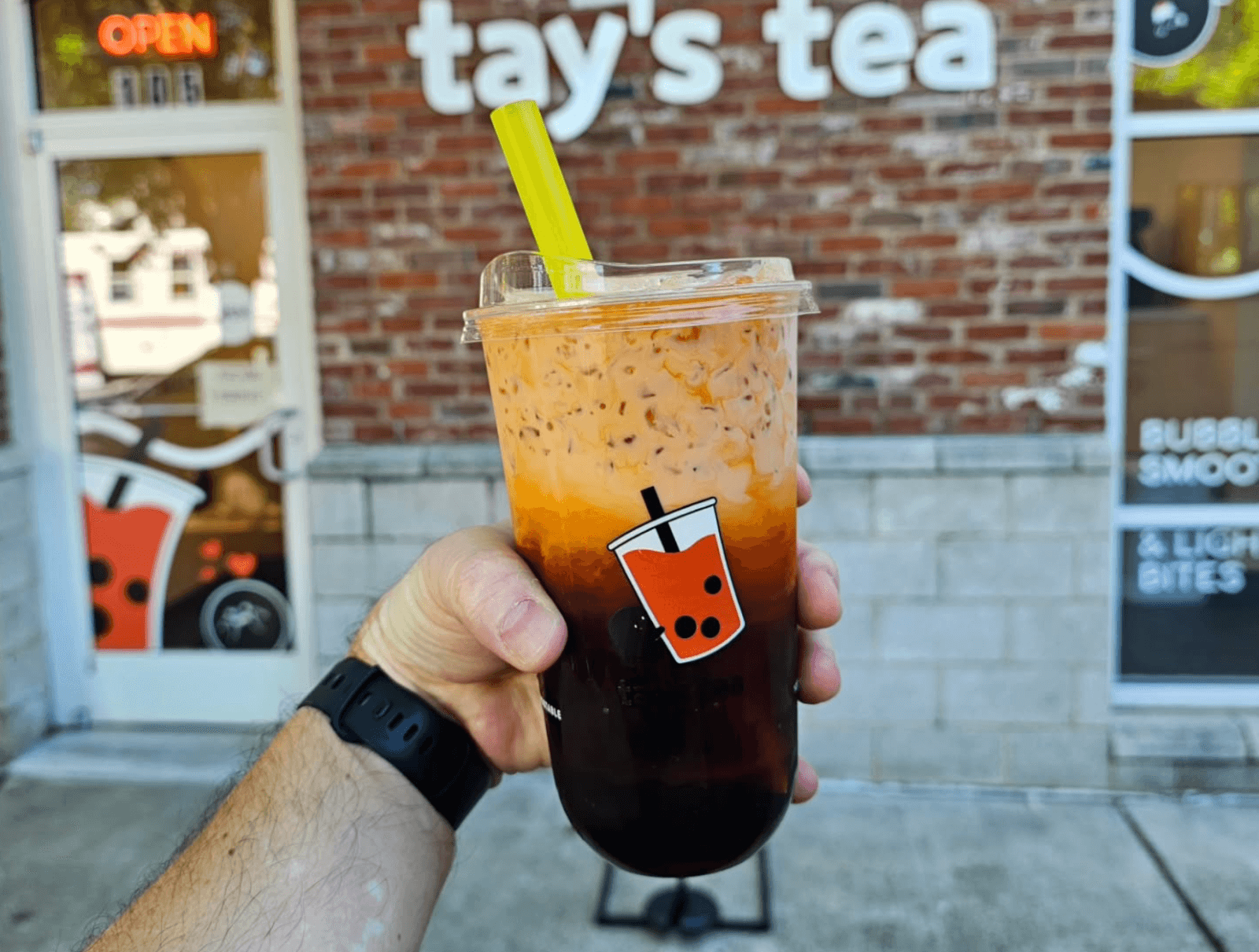 A hand holding a cup of bubble tea with a yellow straw stands before a brick storefront labeled "tay's tea," showcasing fresh new restaurant concepts popular among Birmingham restaurants.