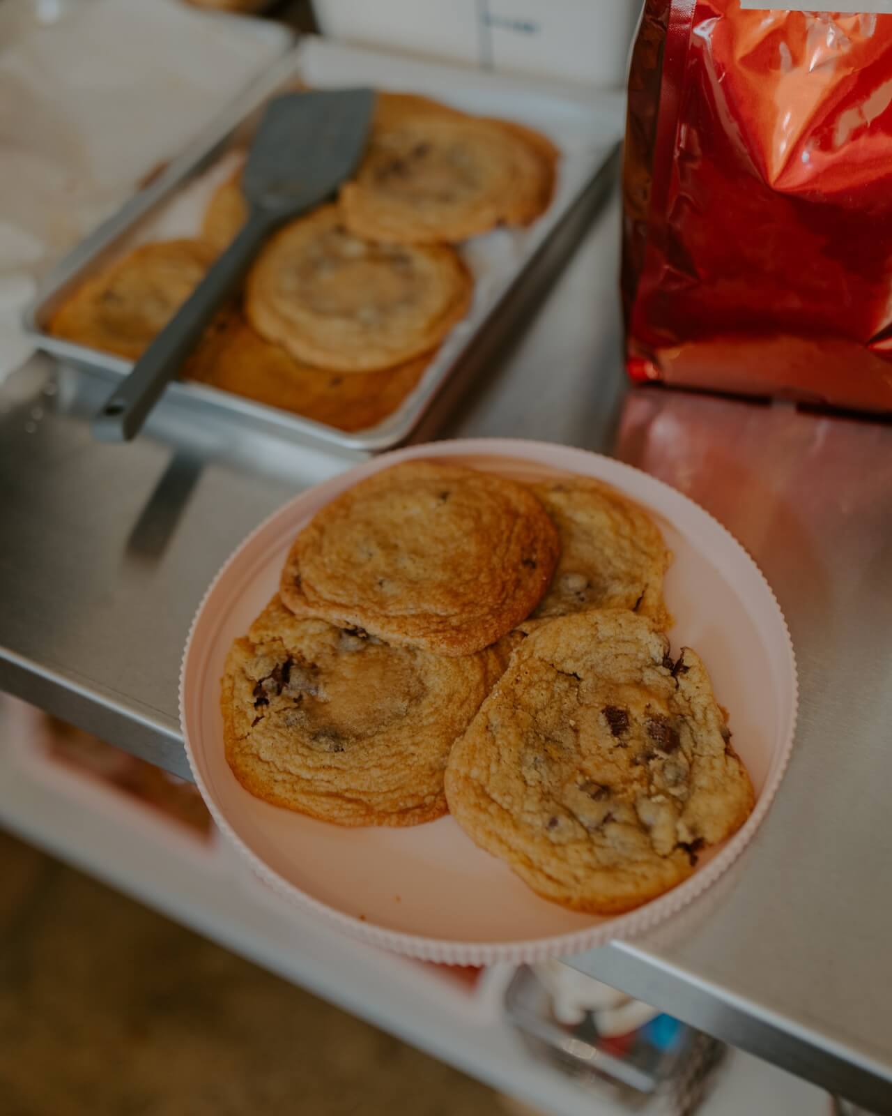 A pink plate with several chocolate chip cookies sits on a metal counter at one of Birmingham’s new restaurants, next to a tray of more cookies and a red bag.